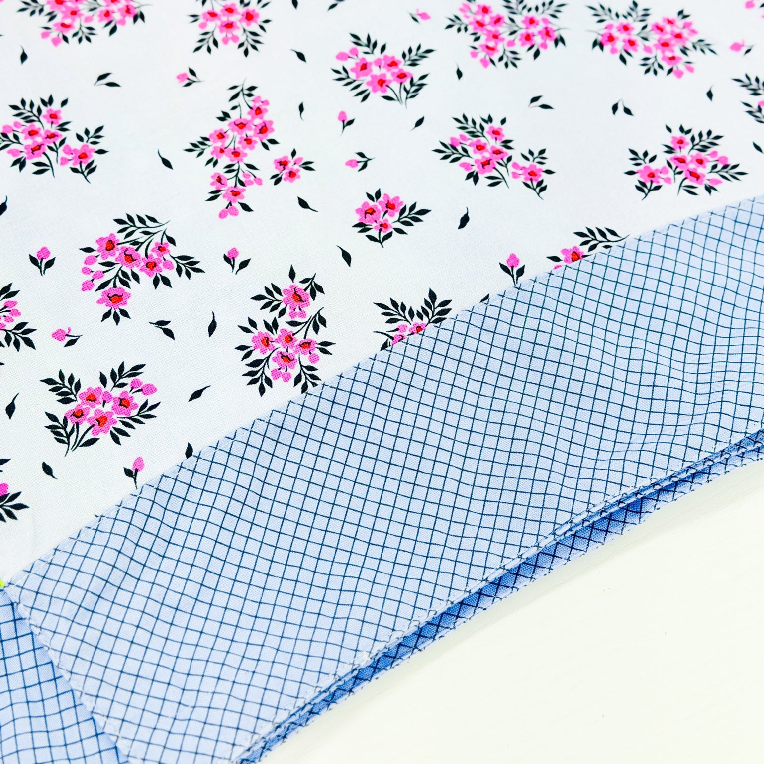 A folded piece of fabric with a white background printed with pink floral clusters and black leaves, layered over a folded piece of light blue and white gingham fabric.