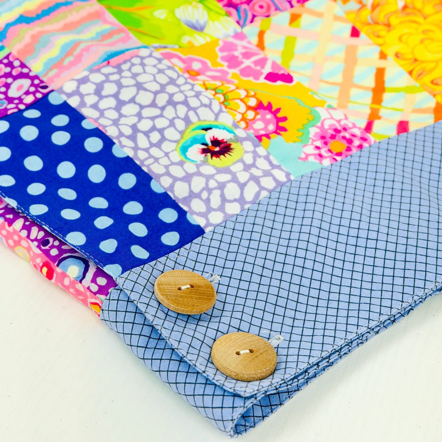 A close-up of a colorful patchwork quilted fabric item, likely a blanket or quilt, featuring a blue gingham folded corner with two wooden buttons.