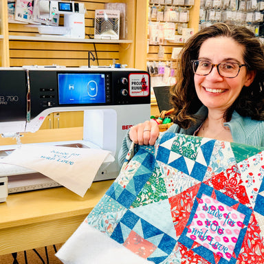 Woman holding a colorful quilt next to a sewing machine in a store setting
