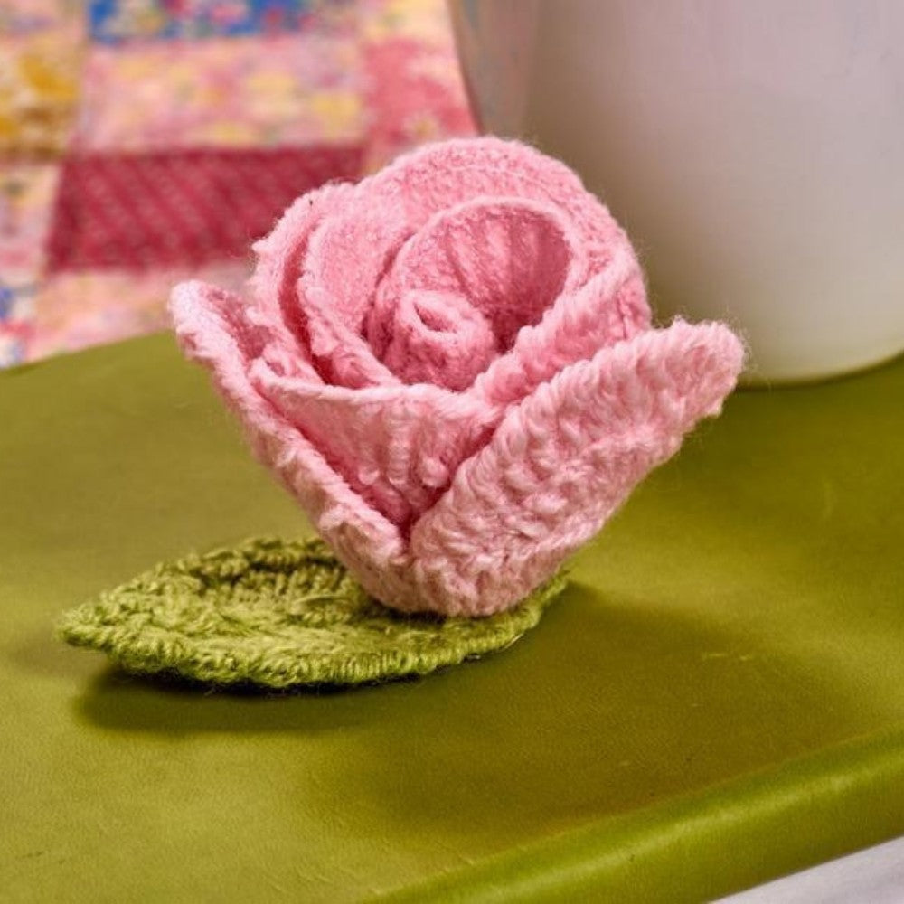Pink knitted flower on a green base with a white cup in the background