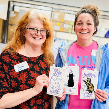 Two women holding cards with cat illustrations in a store setting