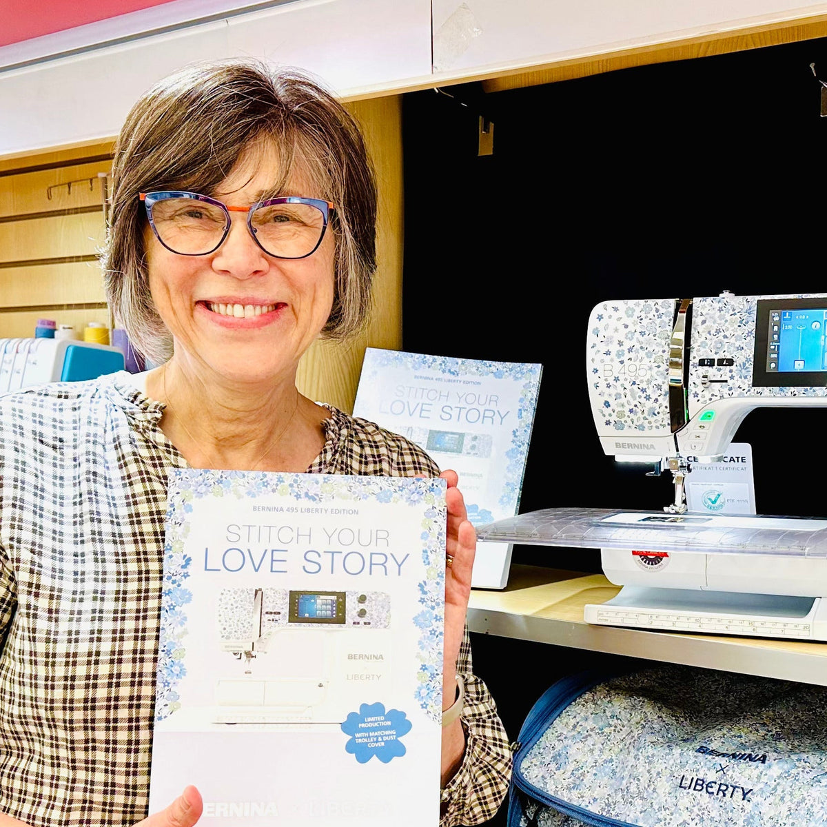 A smiling woman with glasses holds a printed book titled 'Love Story' in a sewing store. Behind her, a white and blue patterned sewing machine sits on a shelf next to a blue patterned suitcase and a trophy.
