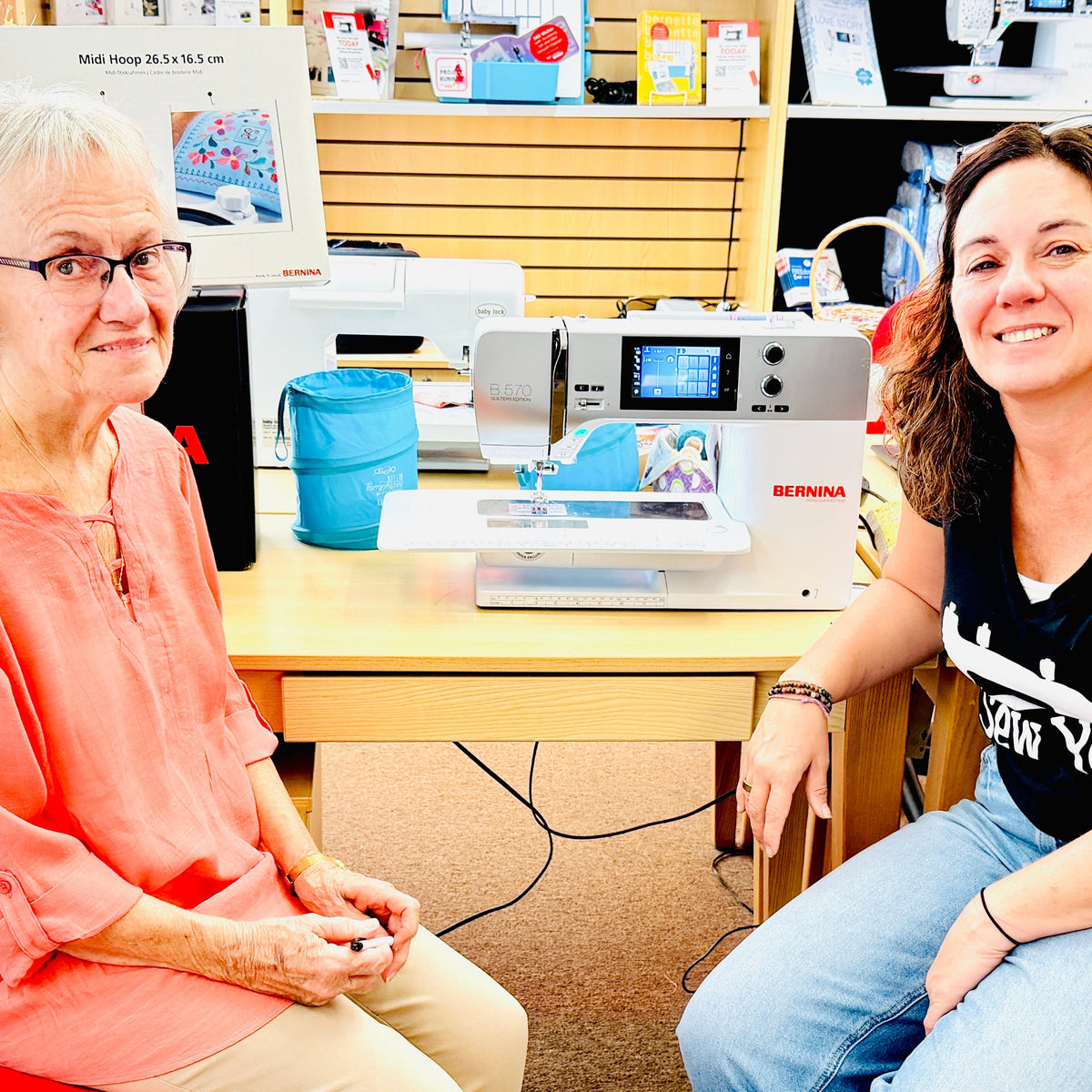 Two women sitting on office chairs in a sewing store, with a red chair on the left and a patterned chair on the right. A white and gray Bernina sewing machine sits on the wooden table between them.