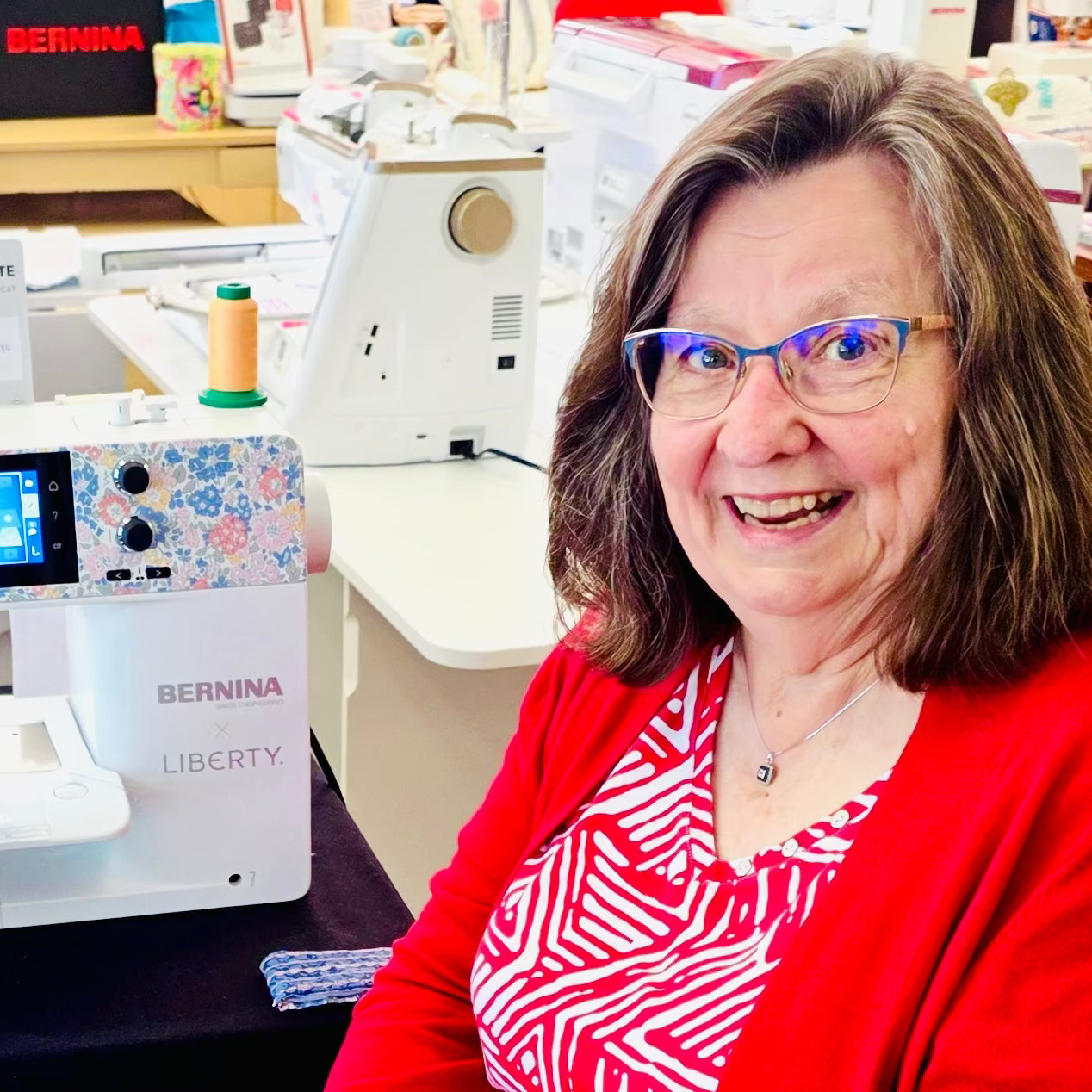 A smiling woman with glasses and brown hair stands next to a BERNINA Liberty embroidery machine in a bright sewing supply store.