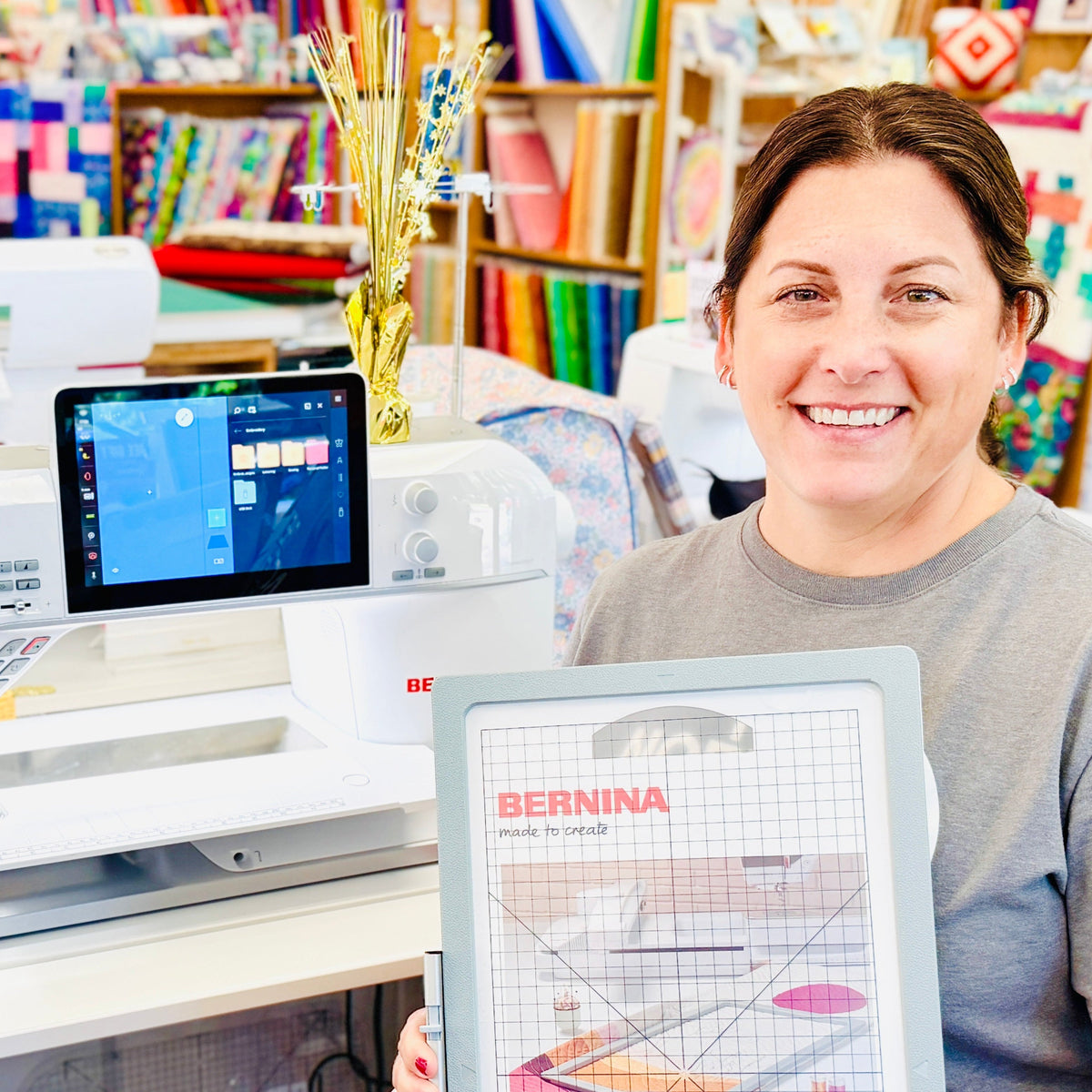 A smiling woman in a gray t-shirt sits in a sewing store holding a BERNINA quilting template. In the background, a white BERNINA sewing machine is visible next to a blue denim tote bag with a colorful heart patch.