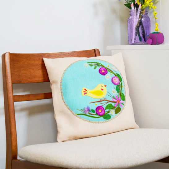 Decorative pillow with a floral design and yellow bird on a chair in a room setting.