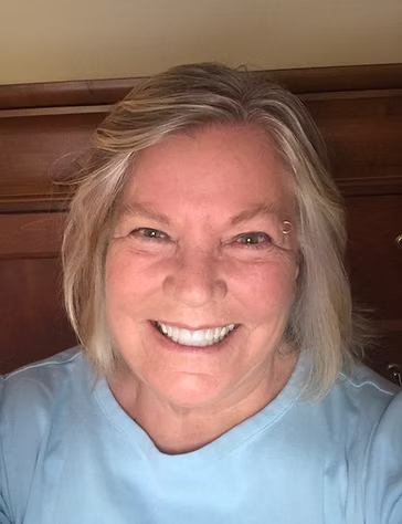 Person with blonde hair wearing a light blue shirt in front of a wooden headboard.