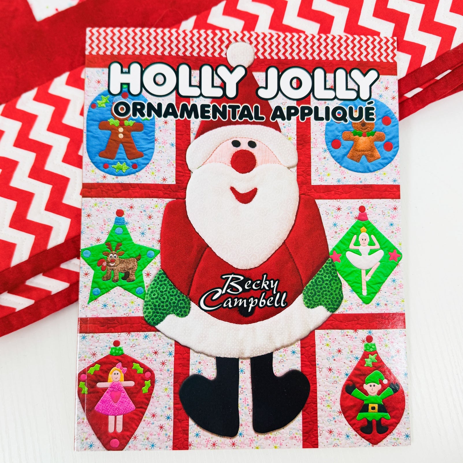 A festive Christmas ornament applique card featuring a central Santa face patch with the text 'HOLLY JOLLY ORNAMENTAL APPLIQUE' and the name 'Recky Campbell' on the chest. The card shows multiple colorful applique designs including gingerbread men, a star ornament, a tree, a girl, and an elf, set against a red and white patterned background with additional red-and-white striped fabric visible in the corners.