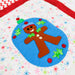 A close-up of a colorful quilted fabric featuring a gingerbread man applique inside a blue circle, surrounded by a white fabric with multicolored snowflake patterns and a red chevron border.