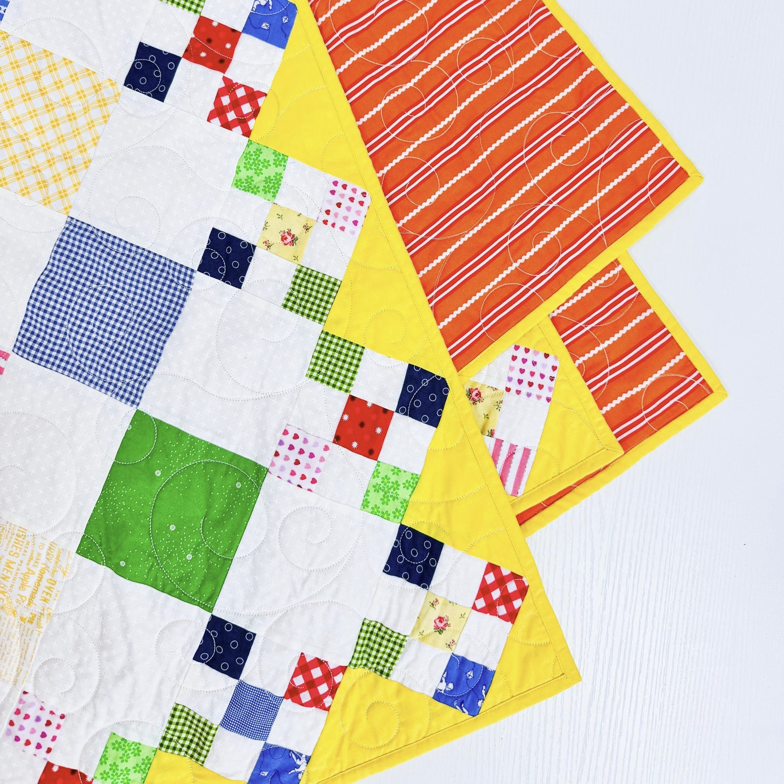 A close-up of a colorful quilted fabric with patchwork squares in white, blue, green, red, yellow, and orange, layered over a bright yellow fabric with red and white stripes, set against a light gray background.