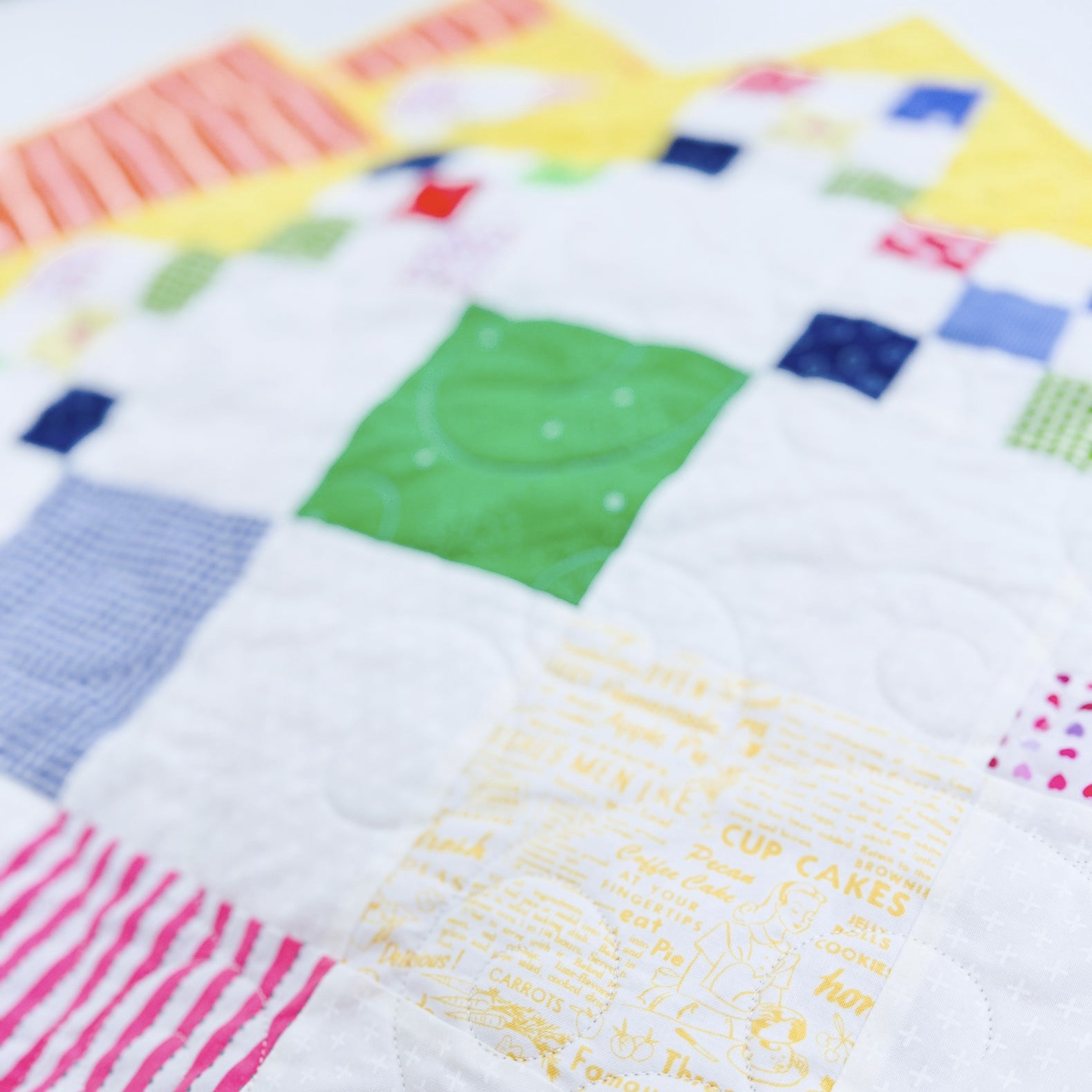 A close-up of a handmade patchwork quilt with various colorful fabric squares, including a green smiley face patch, a pink striped patch, a yellow recipe-print patch, and blue gingham and plaid squares.