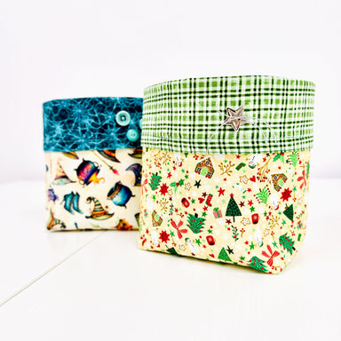 Two fabric storage boxes with buttoned flaps, shown on a white background. The box in front has a green plaid top band and a cream holiday print lower half.