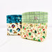 Two fabric storage boxes with buttoned flaps, shown on a white background. The box in front has a green plaid top band and a cream holiday print lower half.