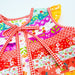 A close-up of a colorful patchwork dress with red gingham trim and a bow, featuring various floral and textured fabric panels in red, yellow, pink, and white.