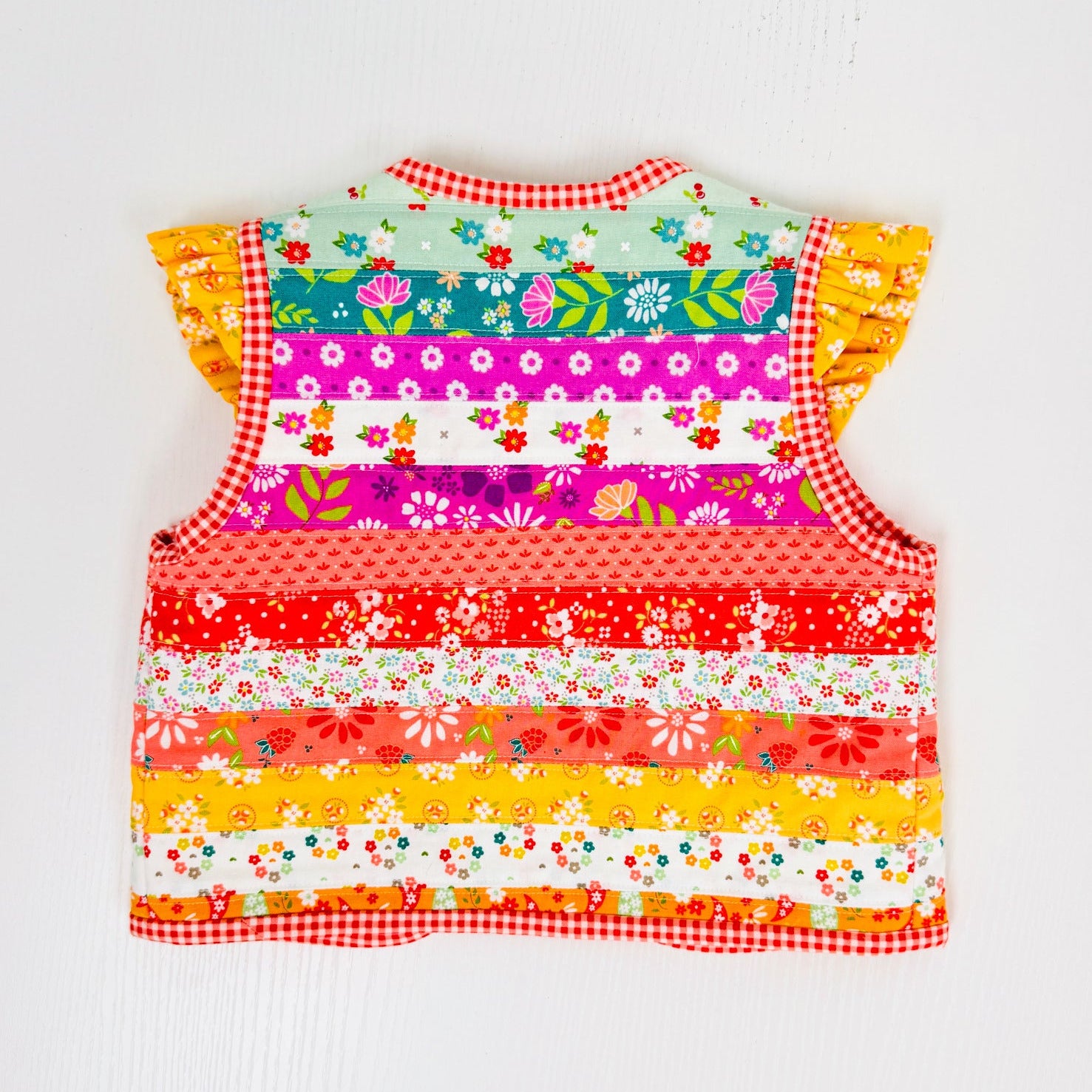 A colorful sleeveless baby vest with ruffle sleeves, featuring a vibrant horizontal striped patchwork design of floral and patterned fabric panels, set against a plain white background.