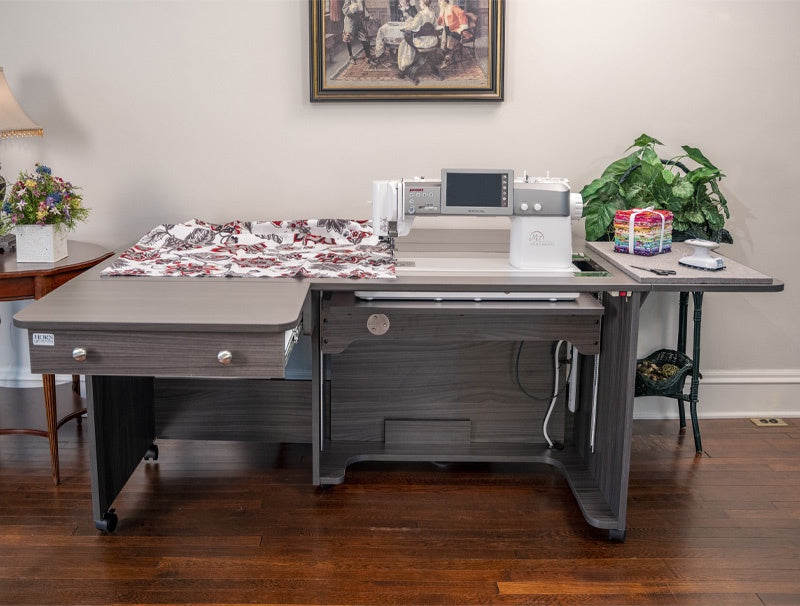 Sewing machine on a large gray table with a floral patterned fabric, in a room with a painting and plants.