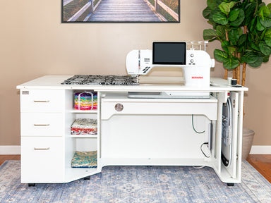 White sewing machine on a dedicated sewing table with storage drawers, in a home setting.