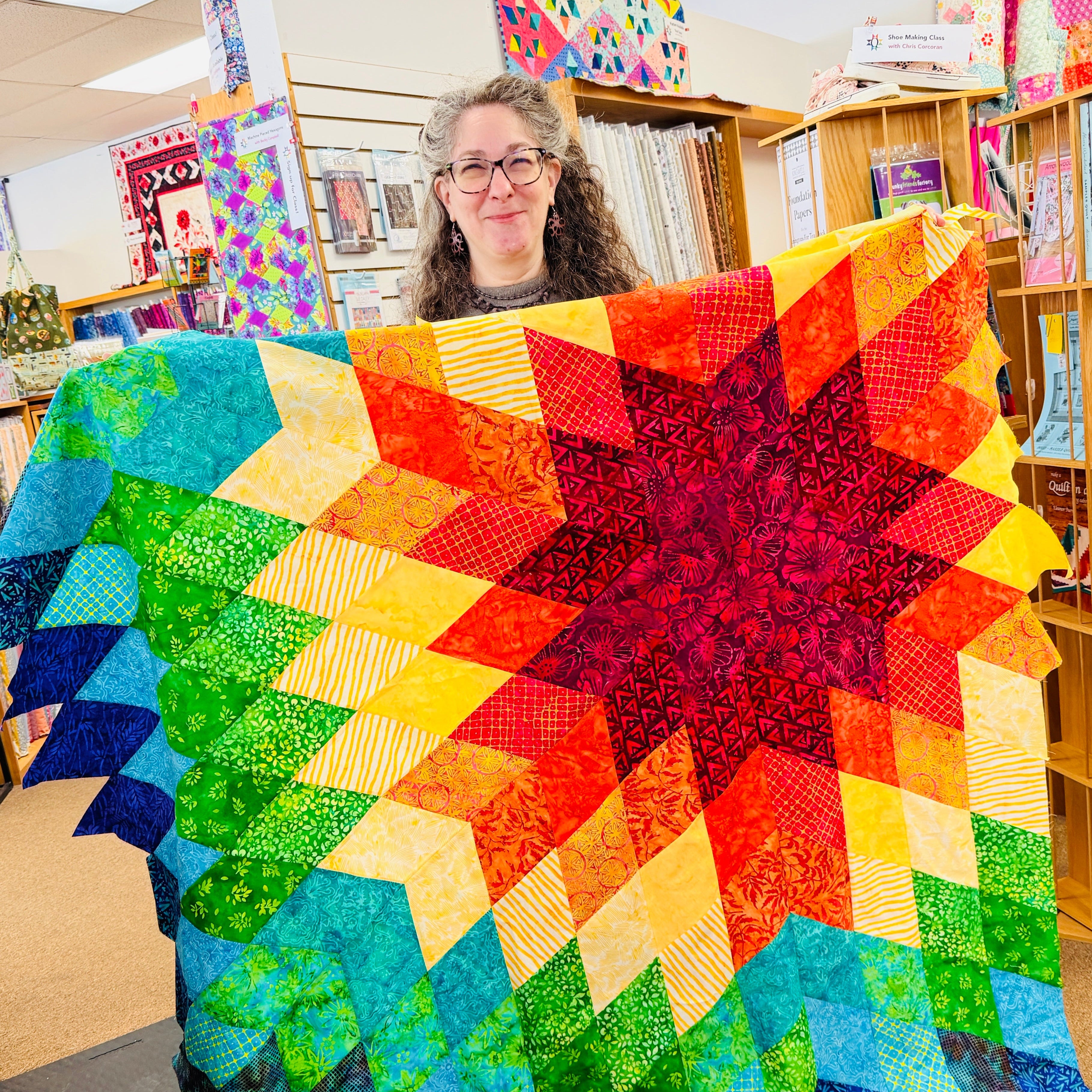 Person holding a colorful star-shaped quilt in a room with bookshelves and other quilts.