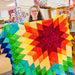 Person holding a colorful star-shaped quilt in a room with bookshelves and other quilts.