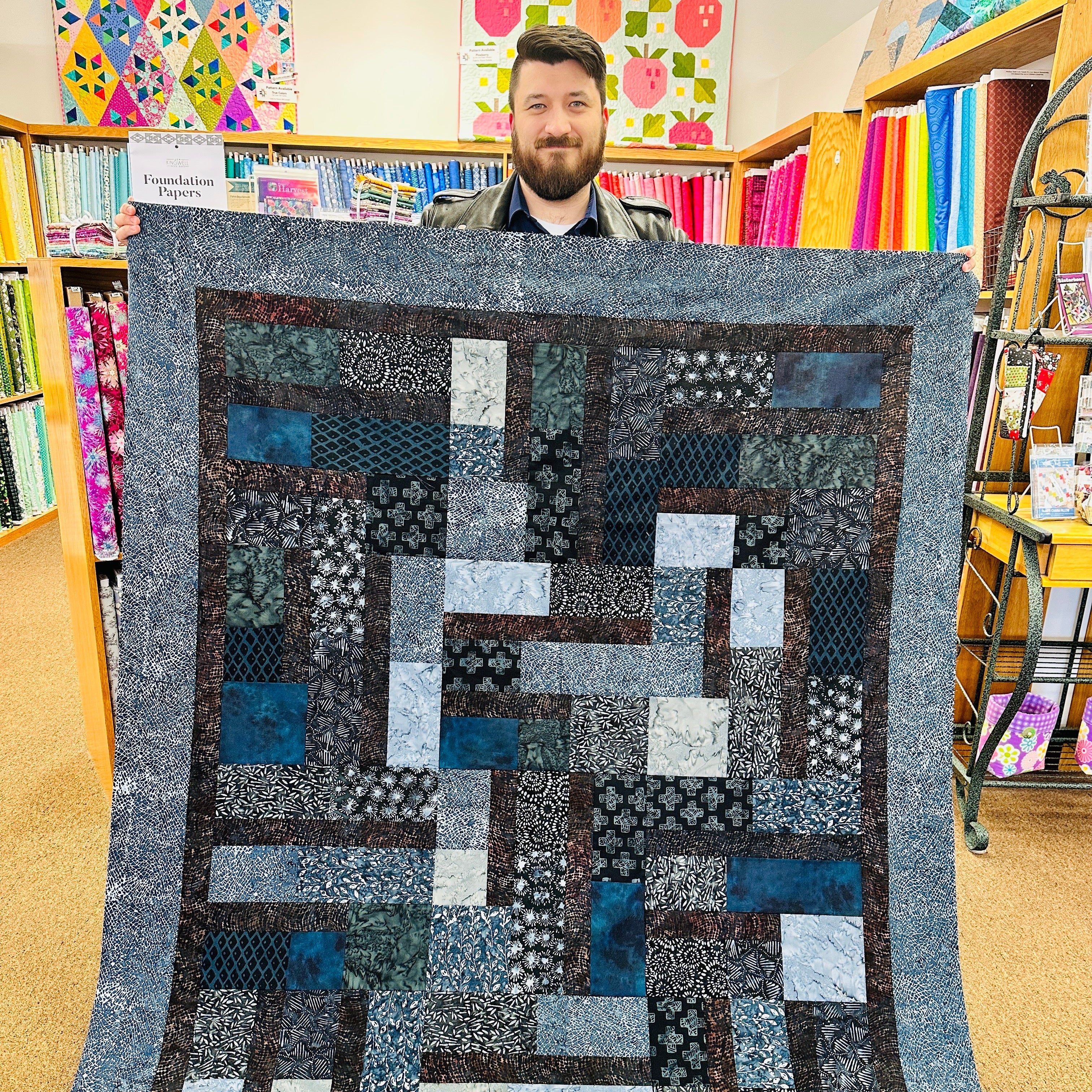 Man holding a large patchwork quilt in a room with bookshelves and colorful decorations.