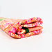 A folded multicolor patterned fabric item, likely a quilt or blanket, featuring a vibrant orange, pink, and white floral design with a small green accent, photographed on a clean white surface.