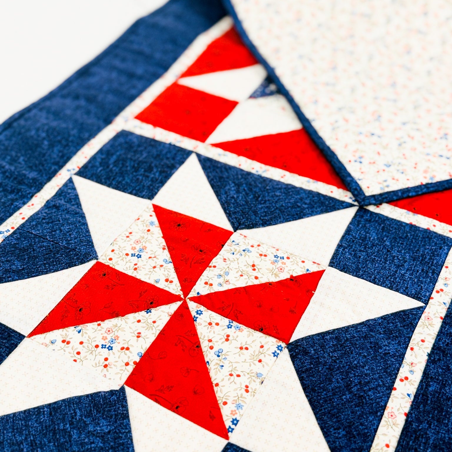 A close-up of a quilted fabric featuring a geometric star pattern in navy blue, white, and red, with a folded corner revealing a light speckled backing.