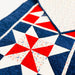 A close-up of a quilted fabric featuring a geometric star pattern in navy blue, white, and red, with a folded corner revealing a light speckled backing.