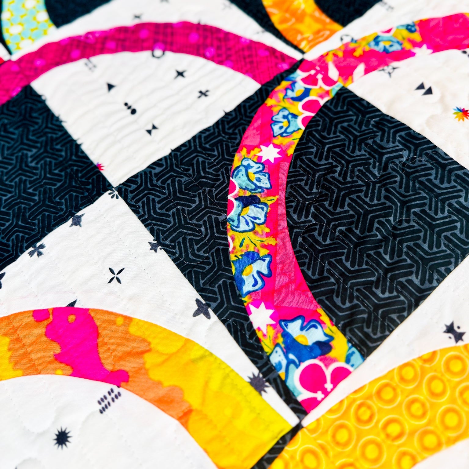 Close-up of a colorful quilted fabric with geometric and circular patchwork designs in black, white, pink, yellow, orange, and blue.