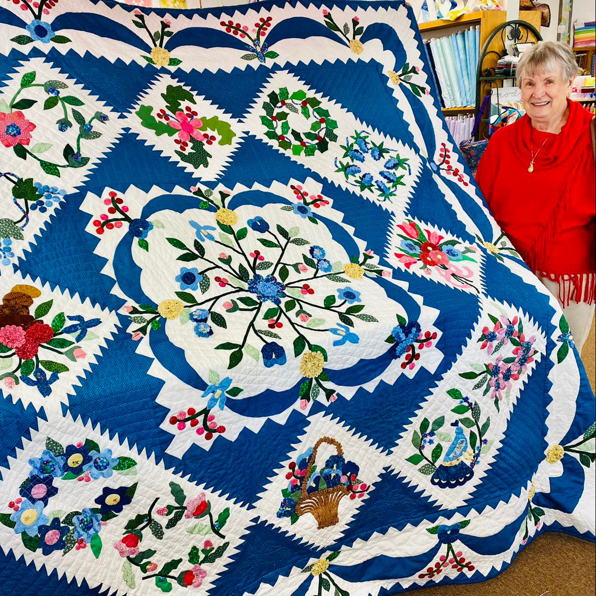 Woman standing next to a large, intricately designed quilt with floral patterns.