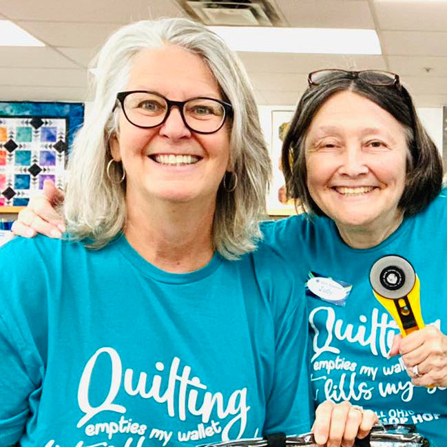 Two women in a classroom setting wearing blue shirts with 'Quilting fills my soul' text, holding quilting-related items.