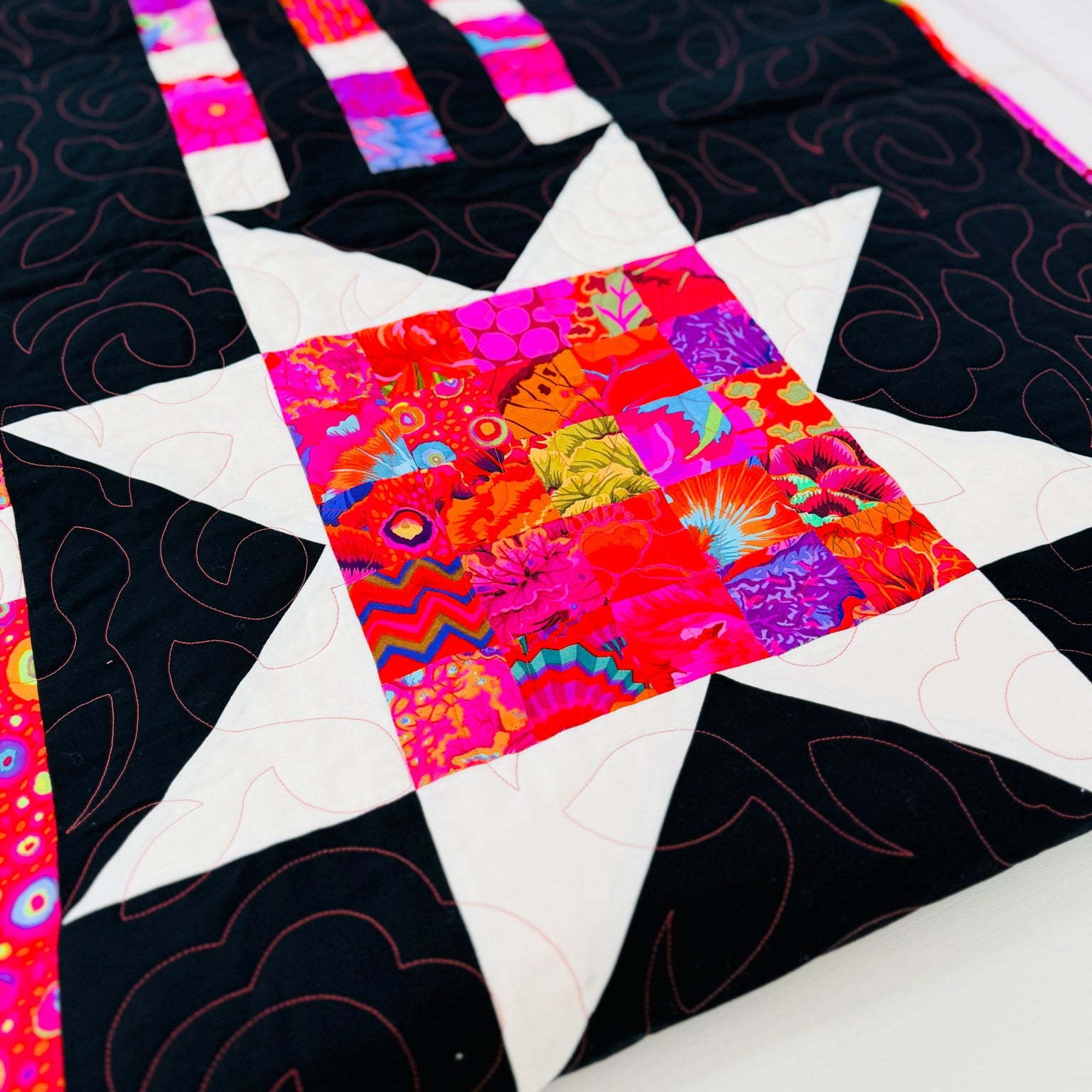 A close-up of a colorful quilted fabric featuring a star pattern with black sections, white points, and a vibrant multicolor patchwork center.