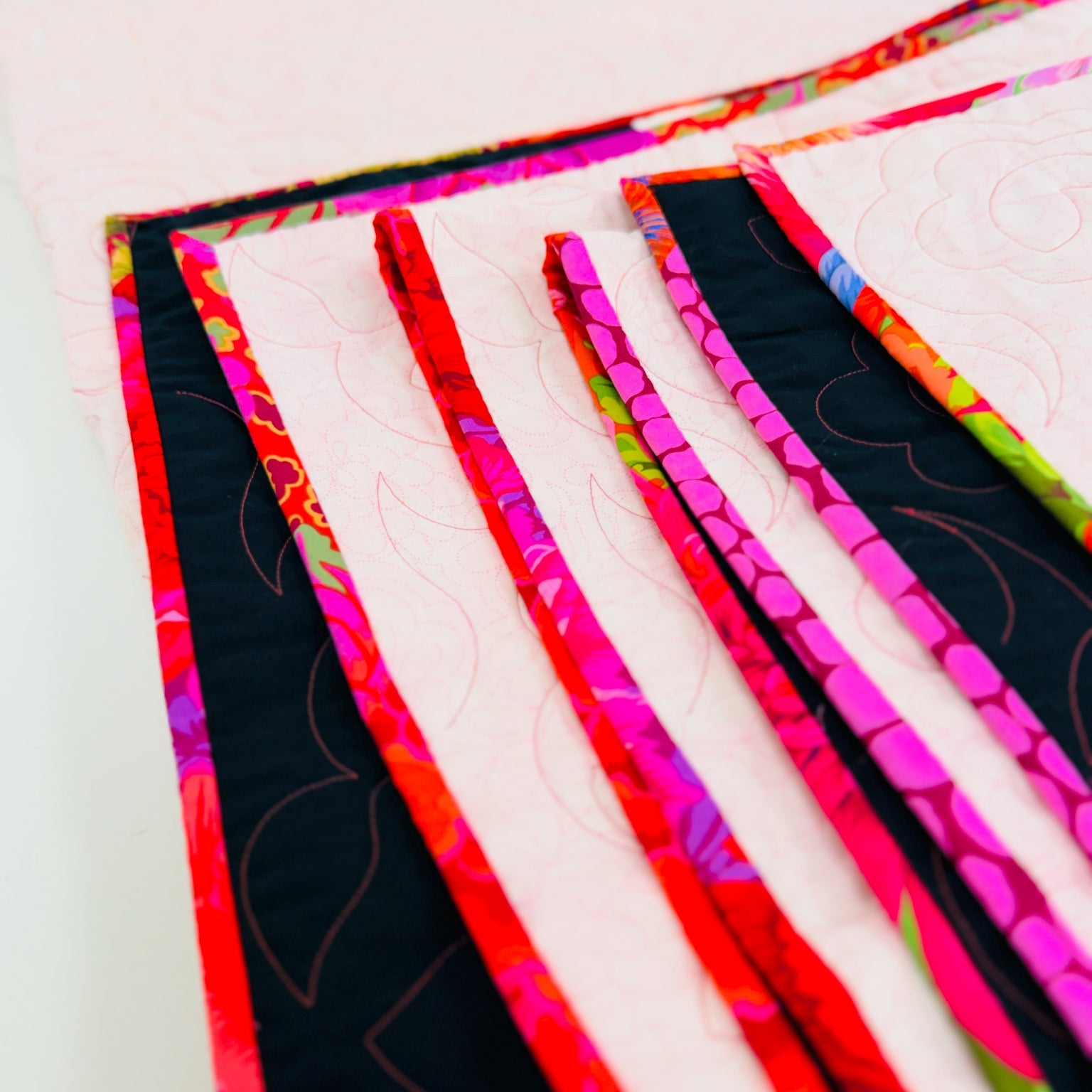 A close-up of a handmade quilted fabric piece with alternating wide light pink panels and narrow black panels trimmed in bright multicolor floral ribbon, photographed on a white background.