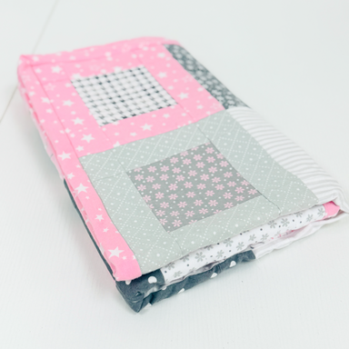 A folded handmade quilted baby blanket with a house design, featuring pink starry fabric, a gray floral patch, and striped details, displayed on a white surface.